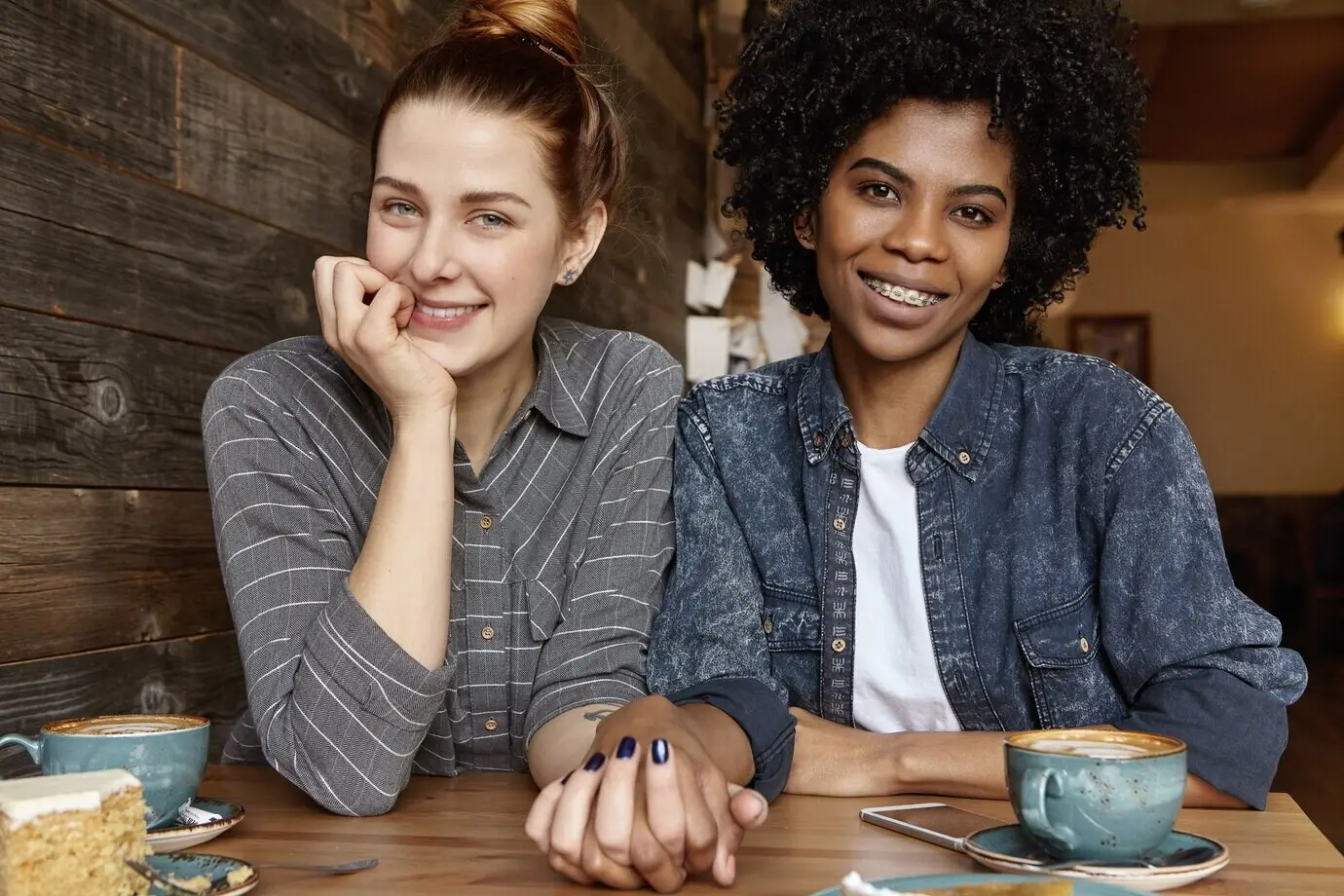 Linda mujer caucásica con el cabello recogido en un moño sostiene la mano de su elegante novia africana durante el almuerzo.